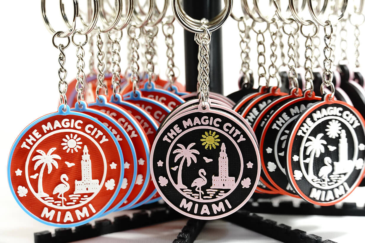 A collection of colorful 3D-printed keychains featuring designs inspired by Miami, including palm trees, a flamingo, and the text 'THE MAGIC CITY MIAMI'.