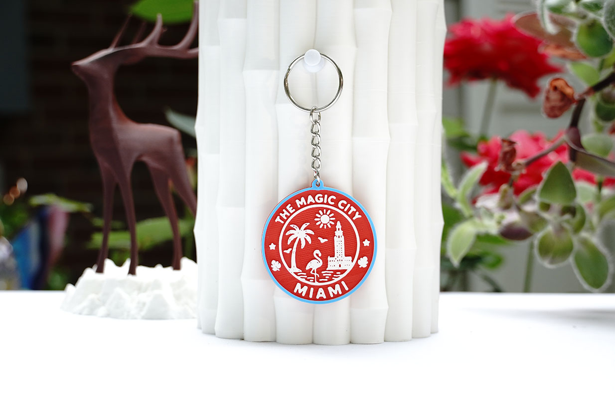 A handcrafted 3D-printed keychain featuring 'The Magic City Miami' design, adorned with palm trees and a flamingo, displayed on a white textured surface with a blurred background of flowers and a decorative deer figurine.