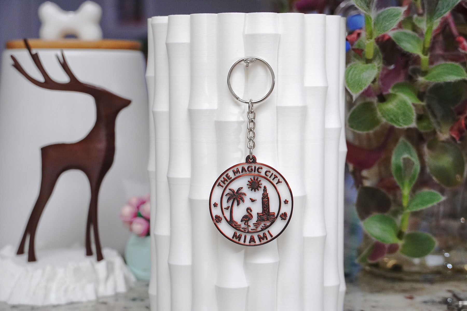 A handcrafted 3D-printed keychain featuring a design inspired by Miami, labeled 'THE MAGIC CITY MIAMI,' displayed next to a decorative white vase and a wooden deer sculpture.